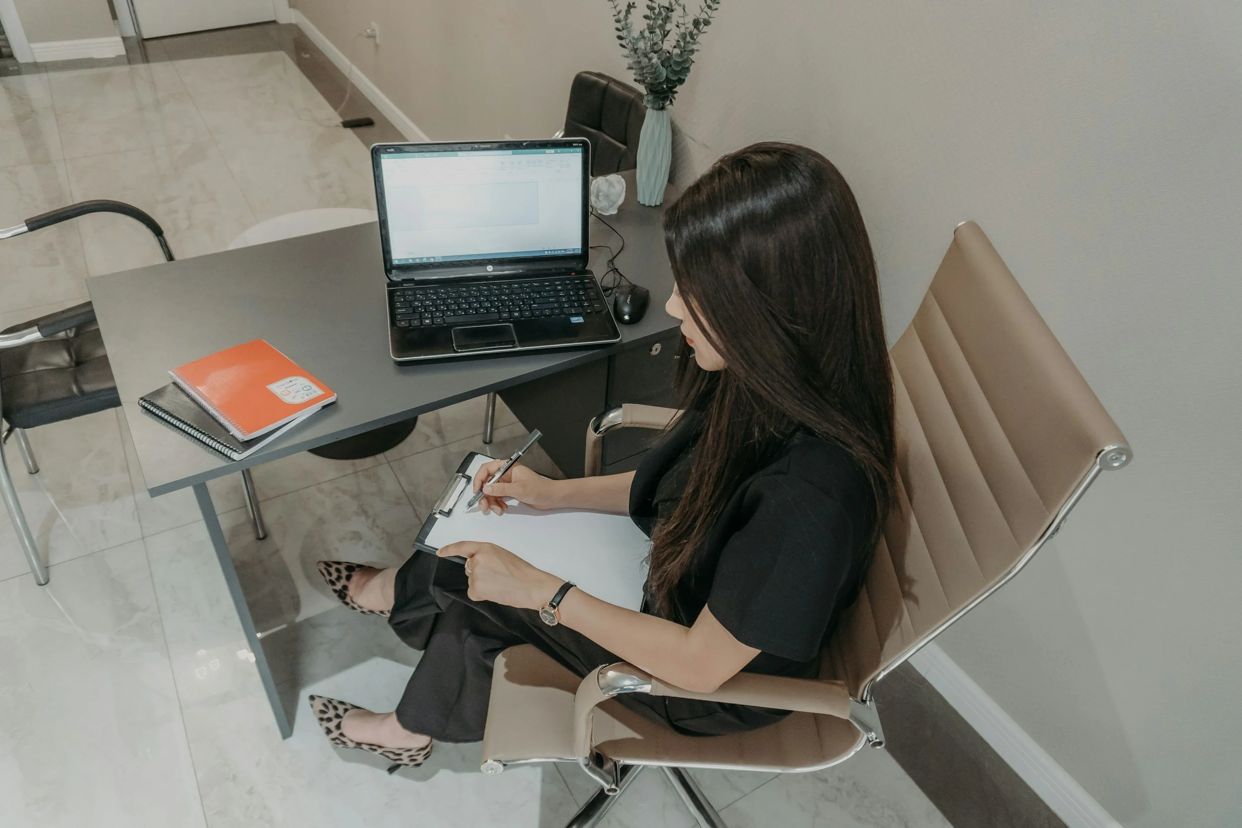 A woman sitting in an office chair at a conference table, taking notes on a notepad, with a laptop and a spiral notebook on the table, and a decorative vase with greenery behind her.