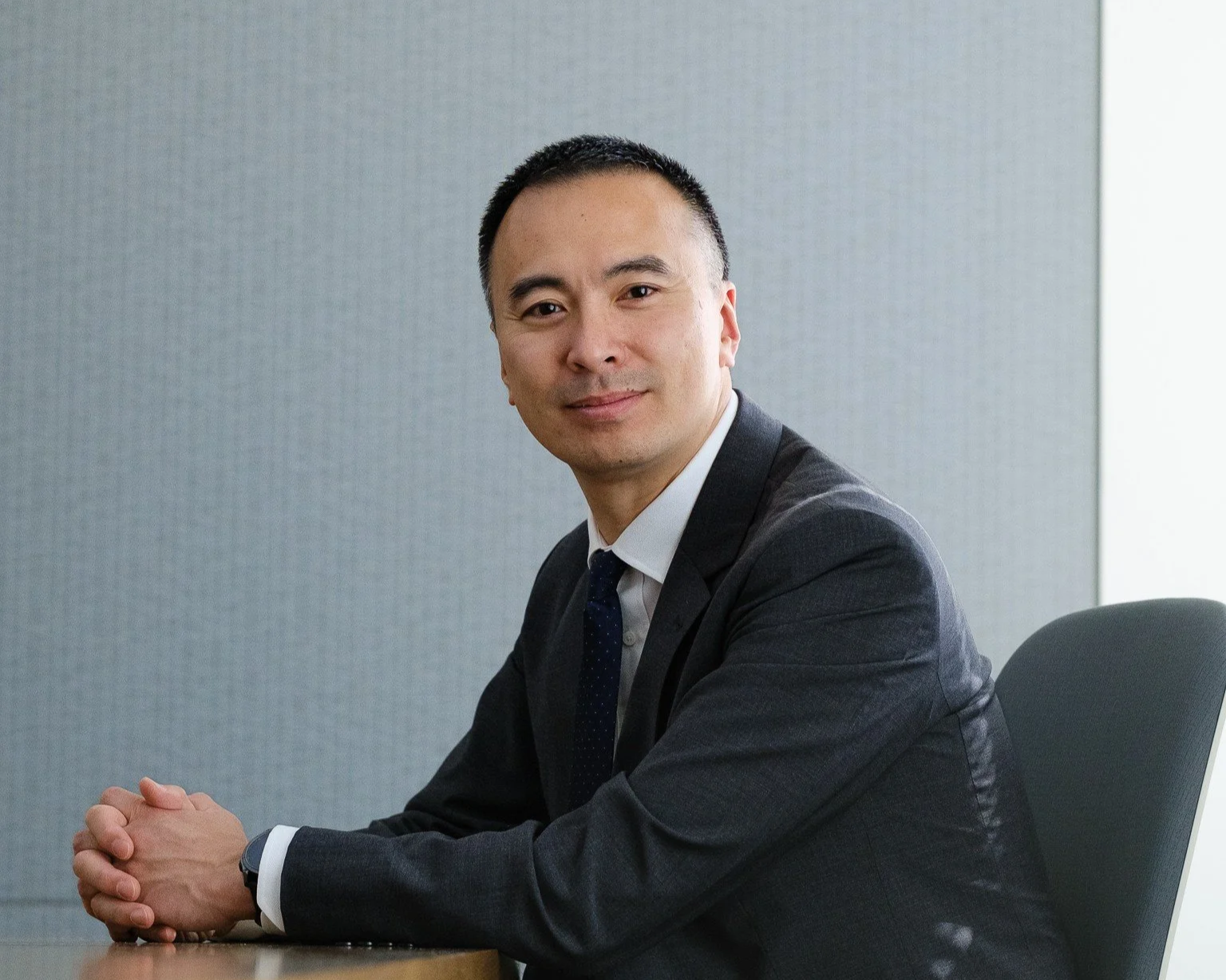 A man in a dark suit and tie sitting at a table in an office setting.