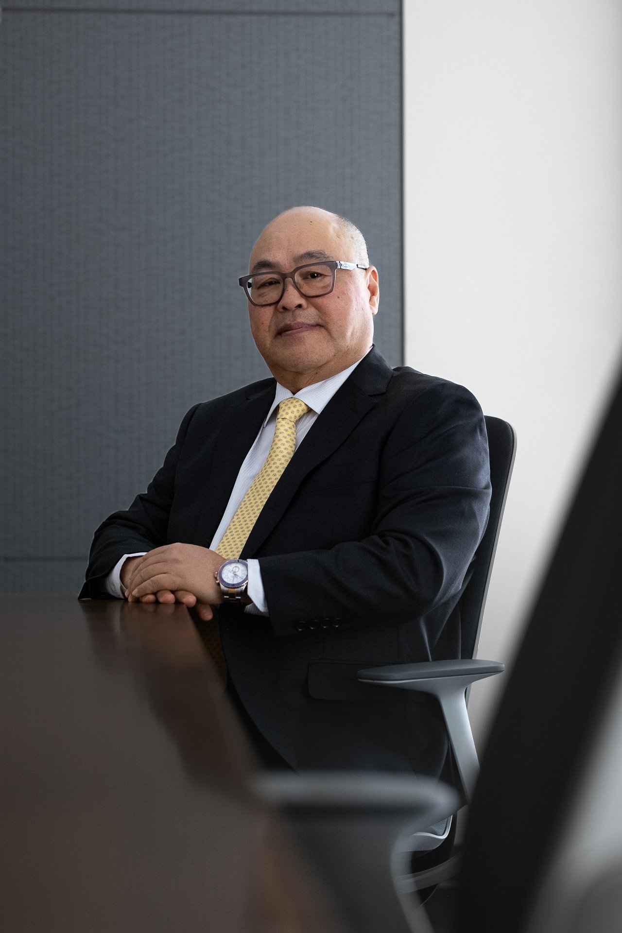 A middle-aged man wearing glasses, a black suit, white shirt, and yellow tie sits at a conference table with his hands clasped. He is in a modern office with a gray and white background.