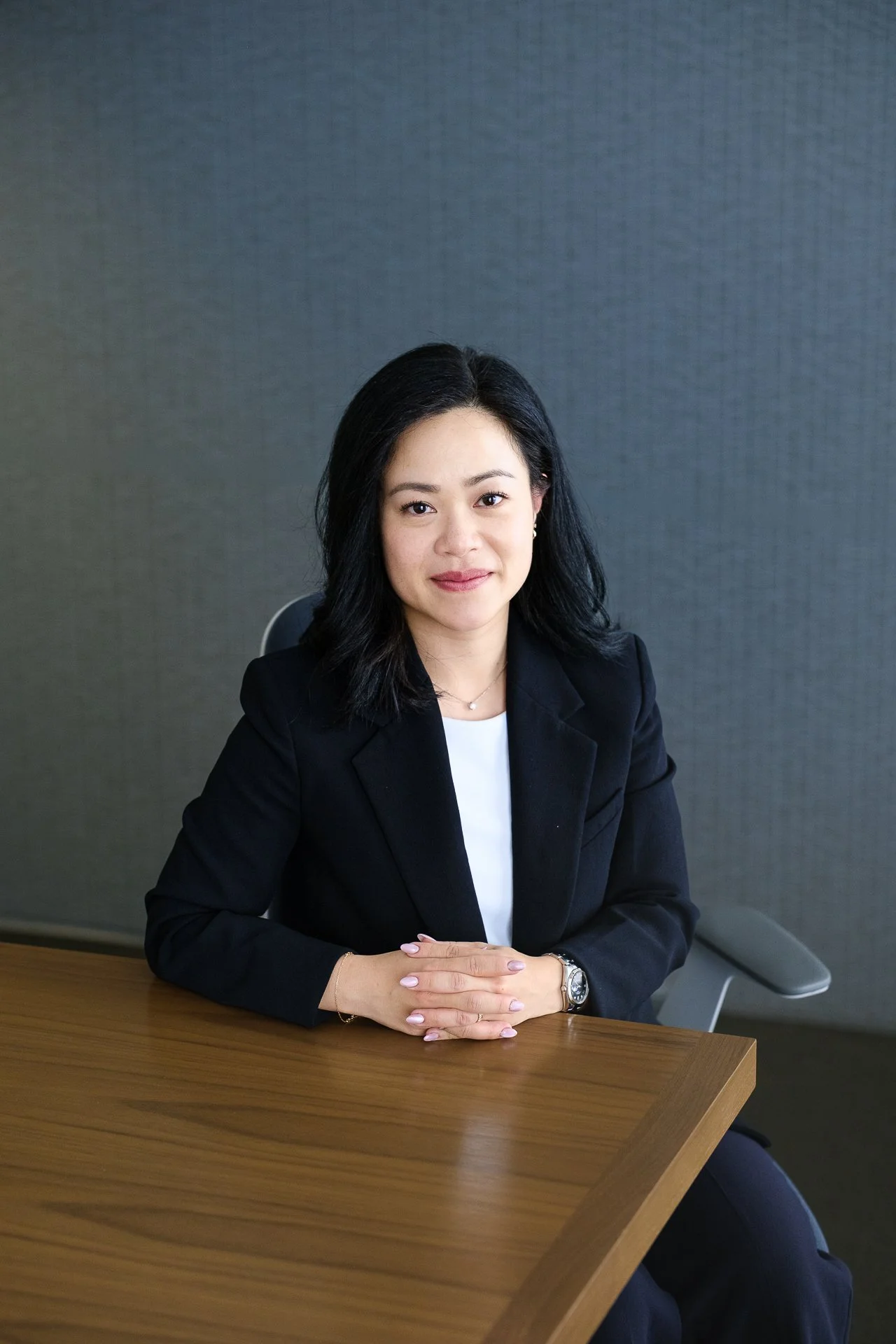 A woman with black hair sitting at a wooden table, wearing a black blazer and white blouse, with her hands clasped on the table, in front of a gray wall background.