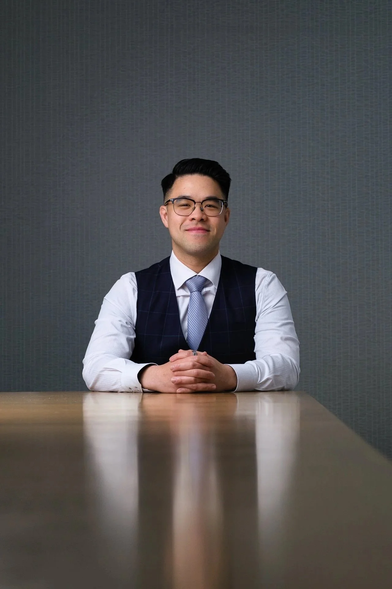 A man in business attire with glasses, sitting at a wooden desk against a plain dark textured wall, looking confidently at the camera.