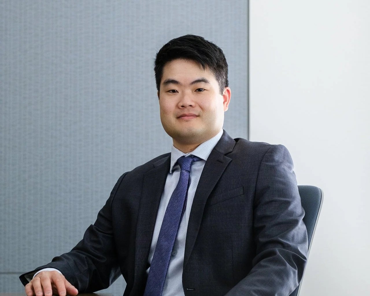 A young Asian man in a dark suit and tie sitting in an office chair in front of a gray and white wall.