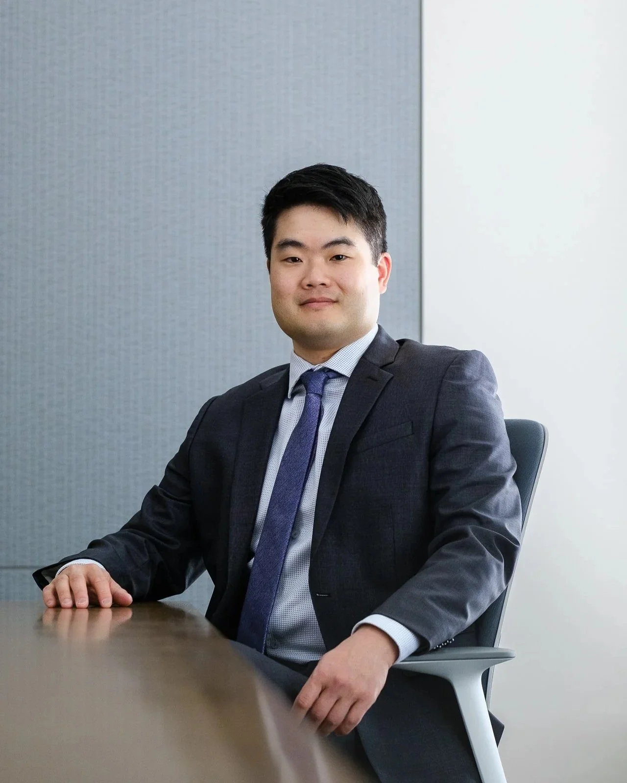 A young Asian man in a dark suit with a light blue shirt and purple tie, sitting confidently in an office chair at a desk, with a neutral expression, against a background of a grey and white office wall.