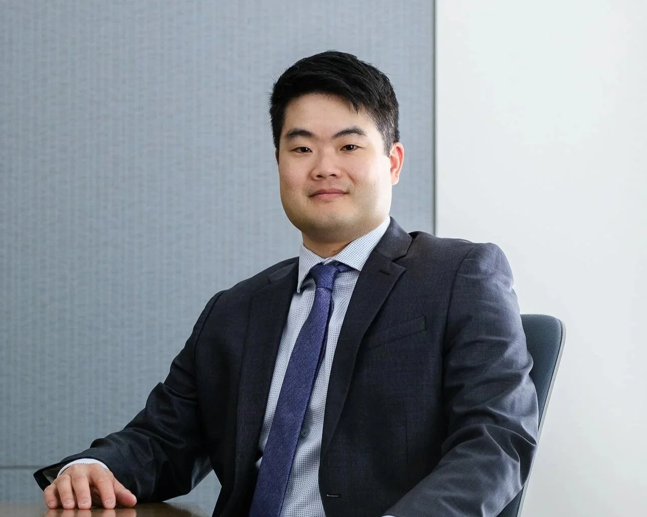 A young man in a dark suit and tie sitting at a table in an office