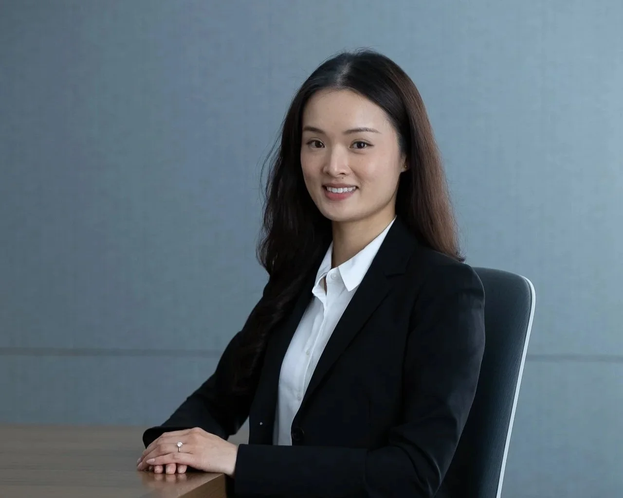 A woman with long dark hair, wearing a black blazer and white blouse, sitting at a desk and smiling at the camera in an office setting.