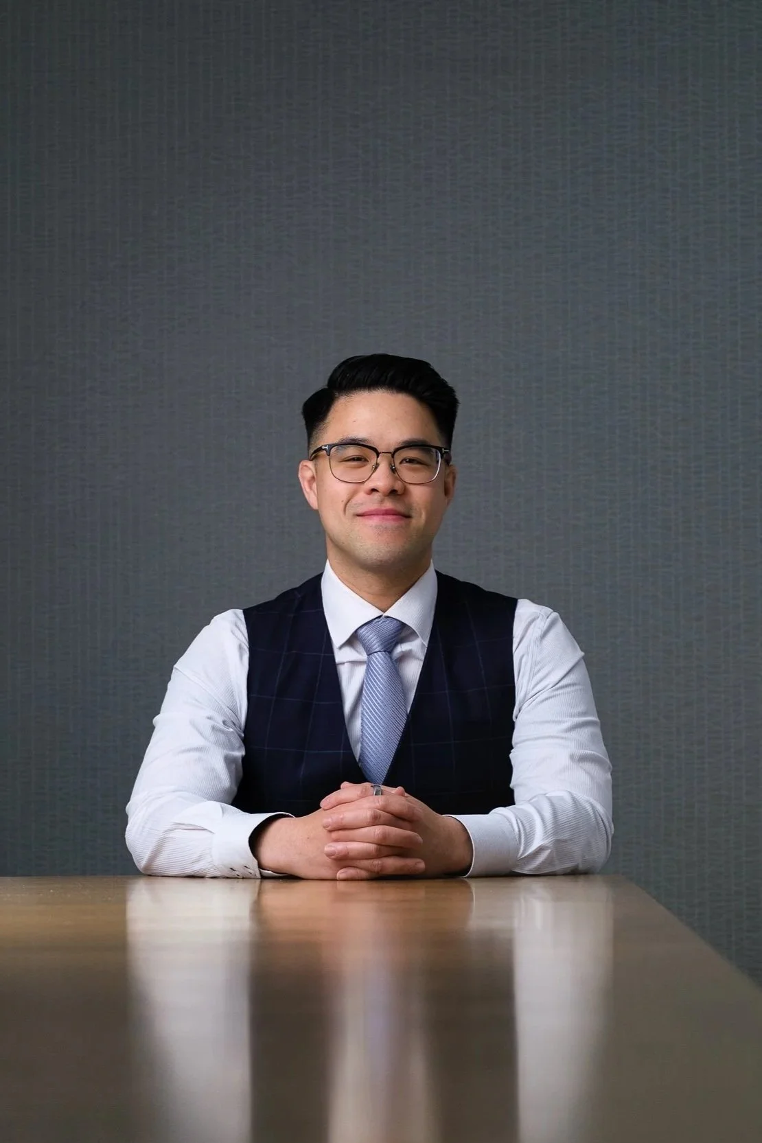 A young man with glasses, wearing a white dress shirt, a blue tie, and a dark vest, sitting at a wooden table with a neutral gray wall background.