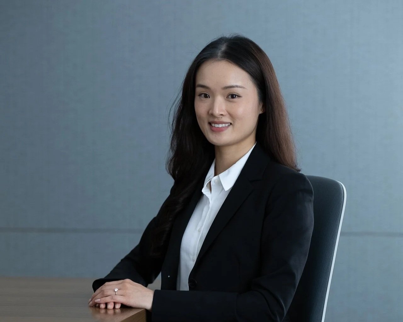 A young woman with long dark hair, wearing a black blazer and white shirt, sitting at a table and smiling at the camera in an office setting.