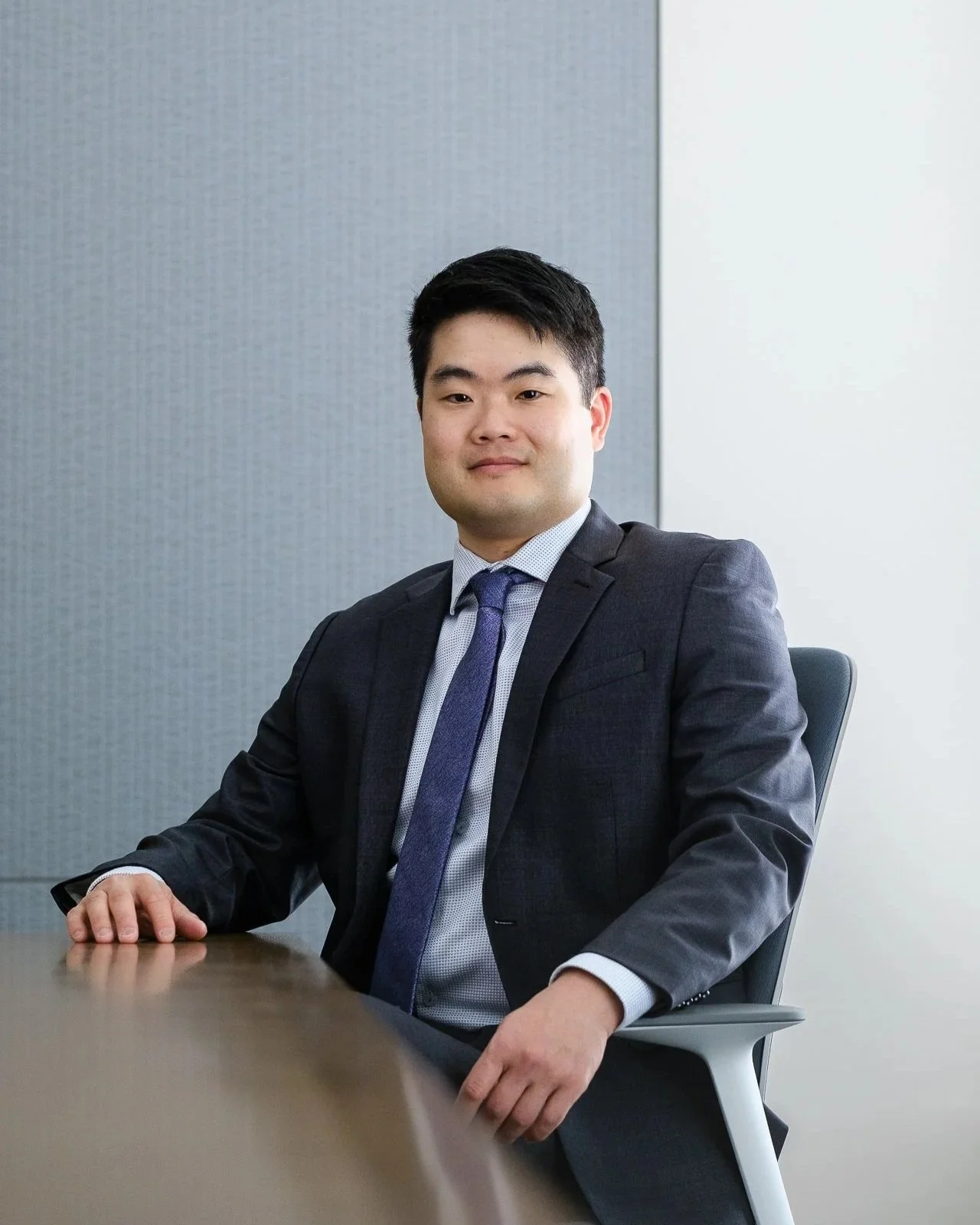A young man in a navy suit with a light blue shirt and dark blue tie sitting at a conference table in an office.