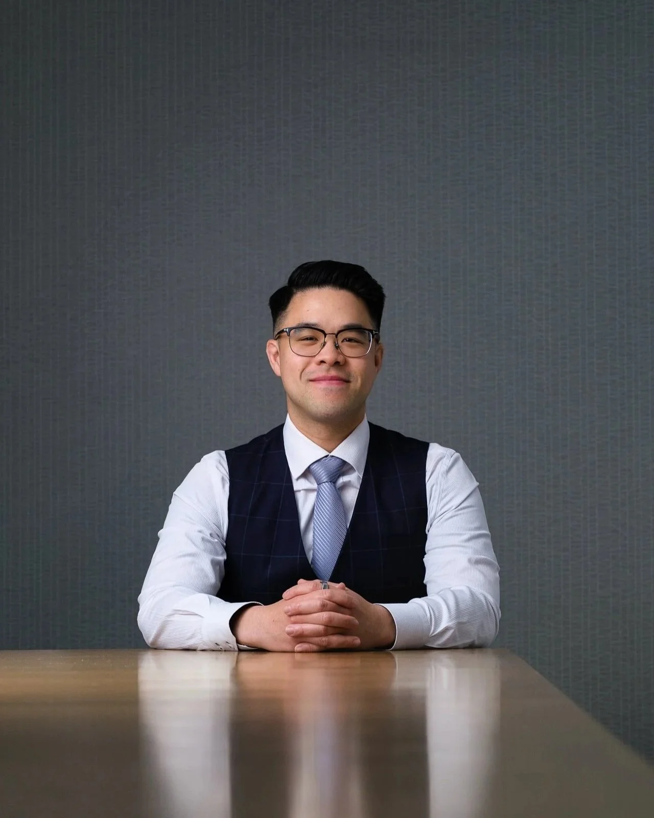 A man in business attire with glasses, sitting at a table, smiling, with hands clasped.