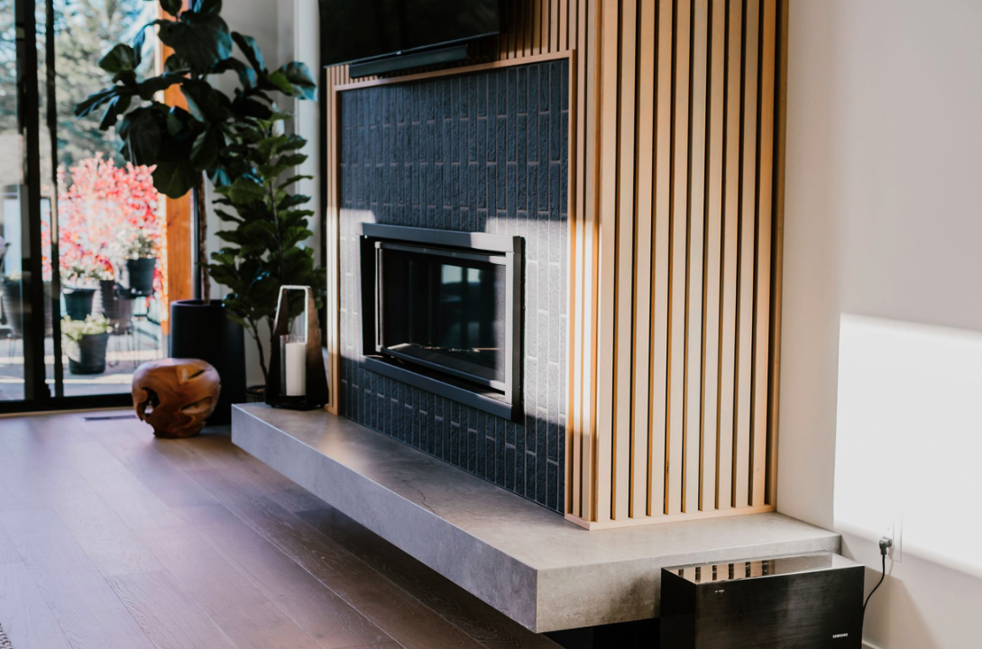 Modern living room corner with a black fireplace embedded in a dark tiled wall, a plant, a decorative wooden object, and a large window with outdoor view.