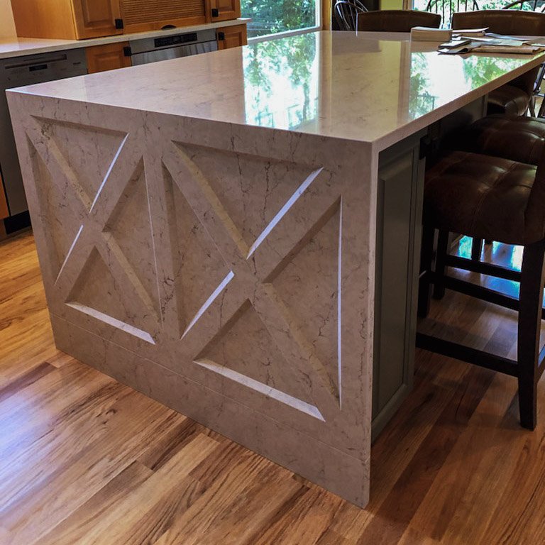 A marble kitchen island with geometric cutouts on its side, situated in a kitchen with wooden floors and a dining area with chairs.