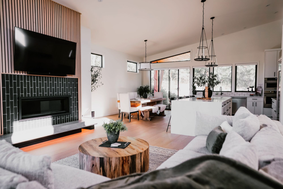 Modern open-concept living room and kitchen with a stone fireplace, flat-screen TV, wooden coffee table, white couch, and large windows letting in natural light.