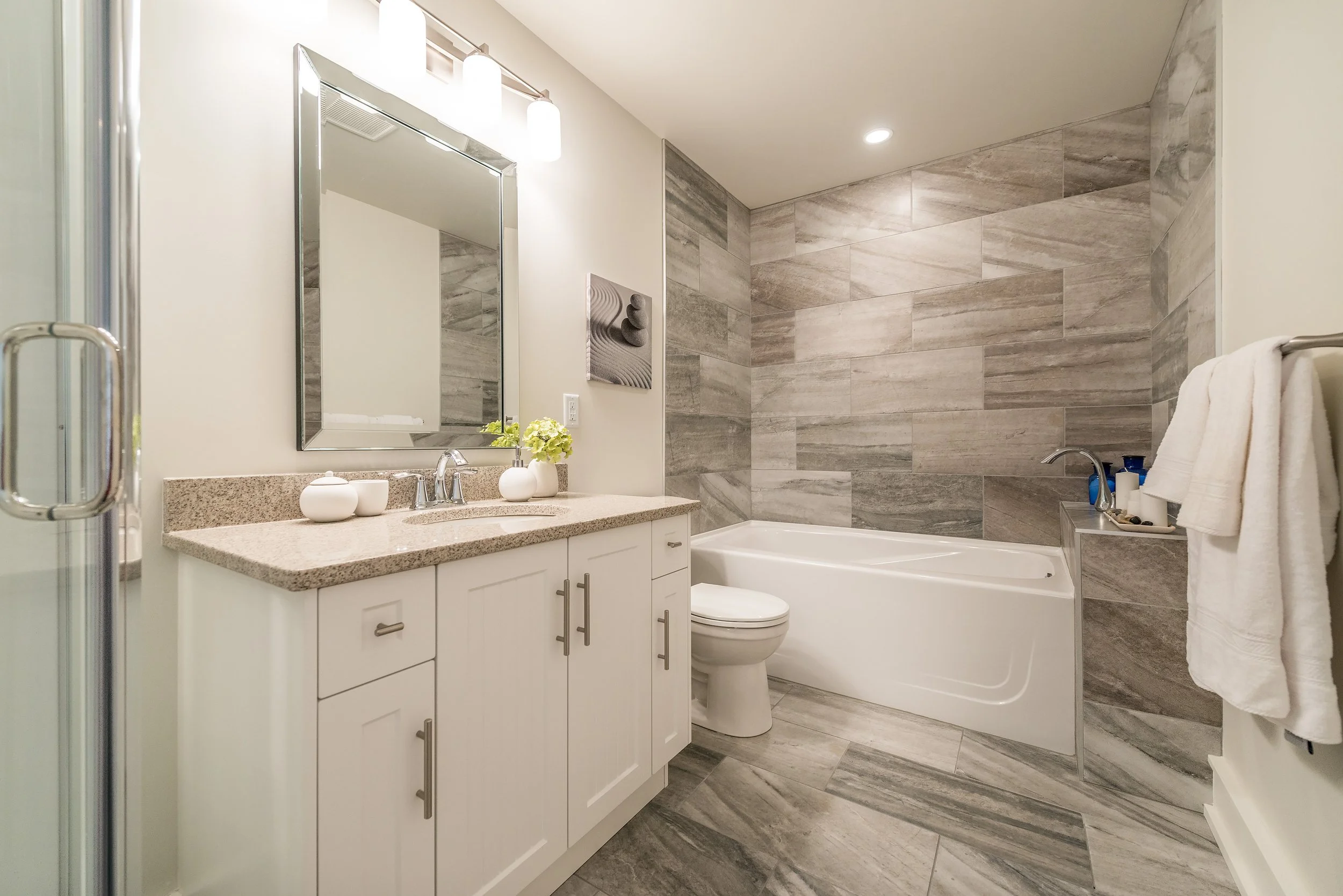 Modern bathroom with a beige granite countertop vanity, mirror, and two white vases, a toilet, a bathtub with beige and gray tiled wall, and a towel rack with a white towel.