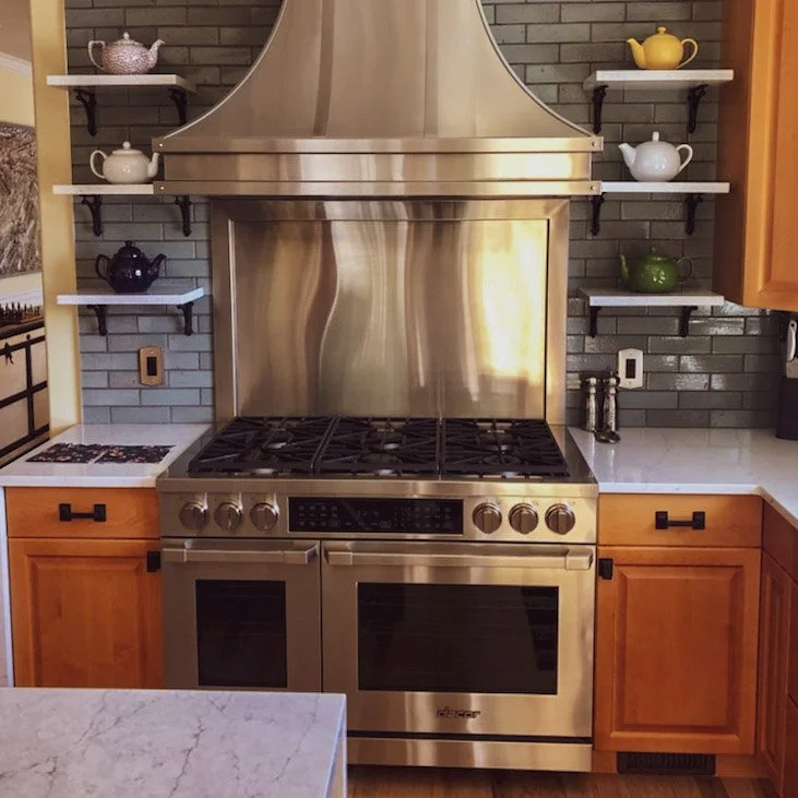A stainless steel gas stove with an oven in a kitchen, with a stainless steel range hood above. There are wooden cabinets on each side and white marble countertops. Two open shelves behind the stove hold teapots in various colors and designs, set aga