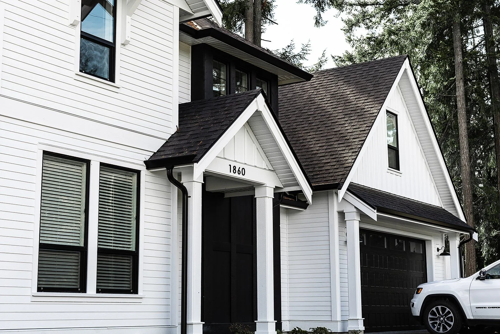 Front view of a modern white house with black accents, multiple windows, and a garage with a white vehicle parked outside, surrounded by tall trees.