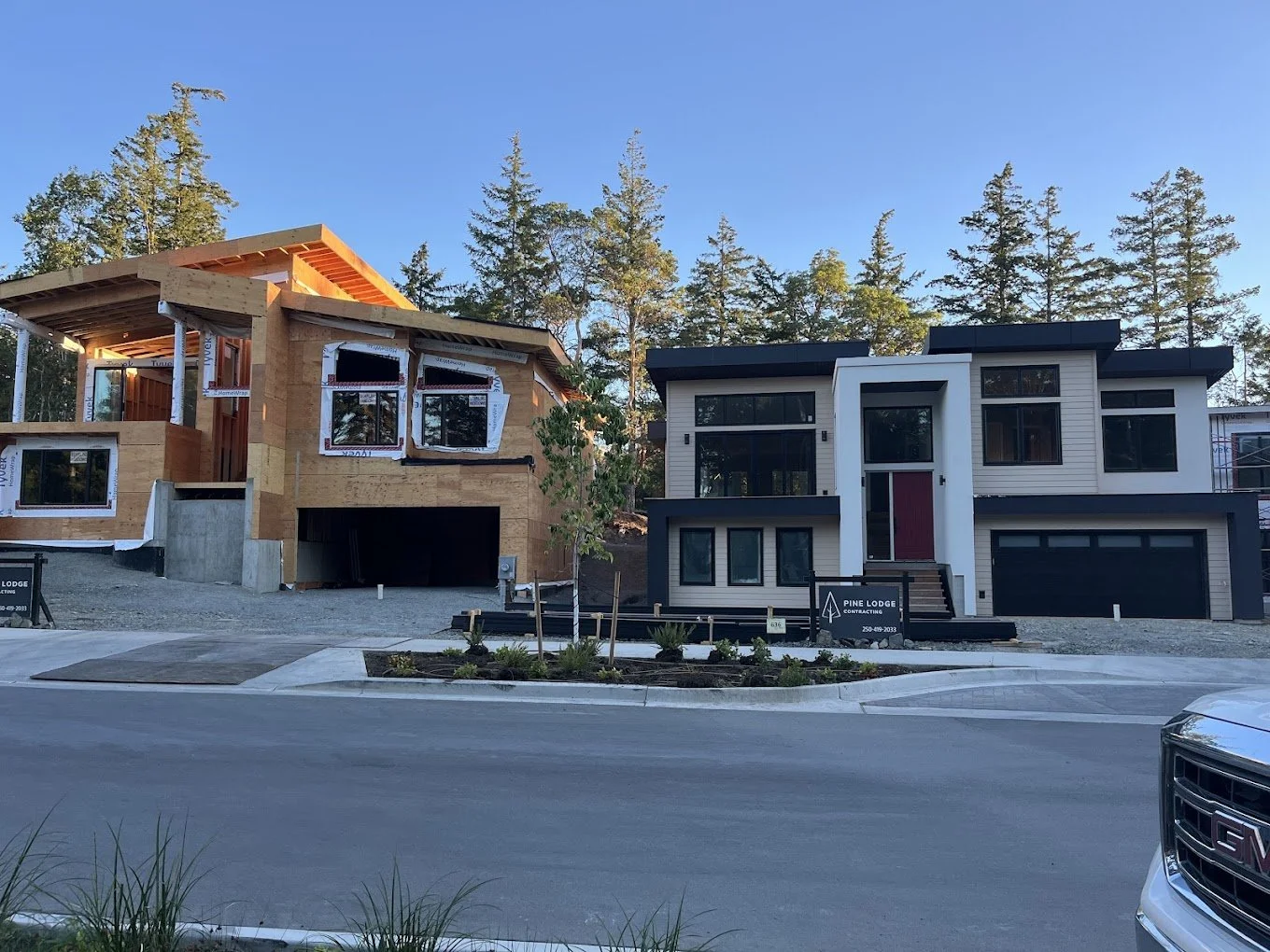 Two modern houses under construction, one partially built with exposed wood framing and the other completed with white and dark gray exterior, situated in a wooded area with tall trees in the background.