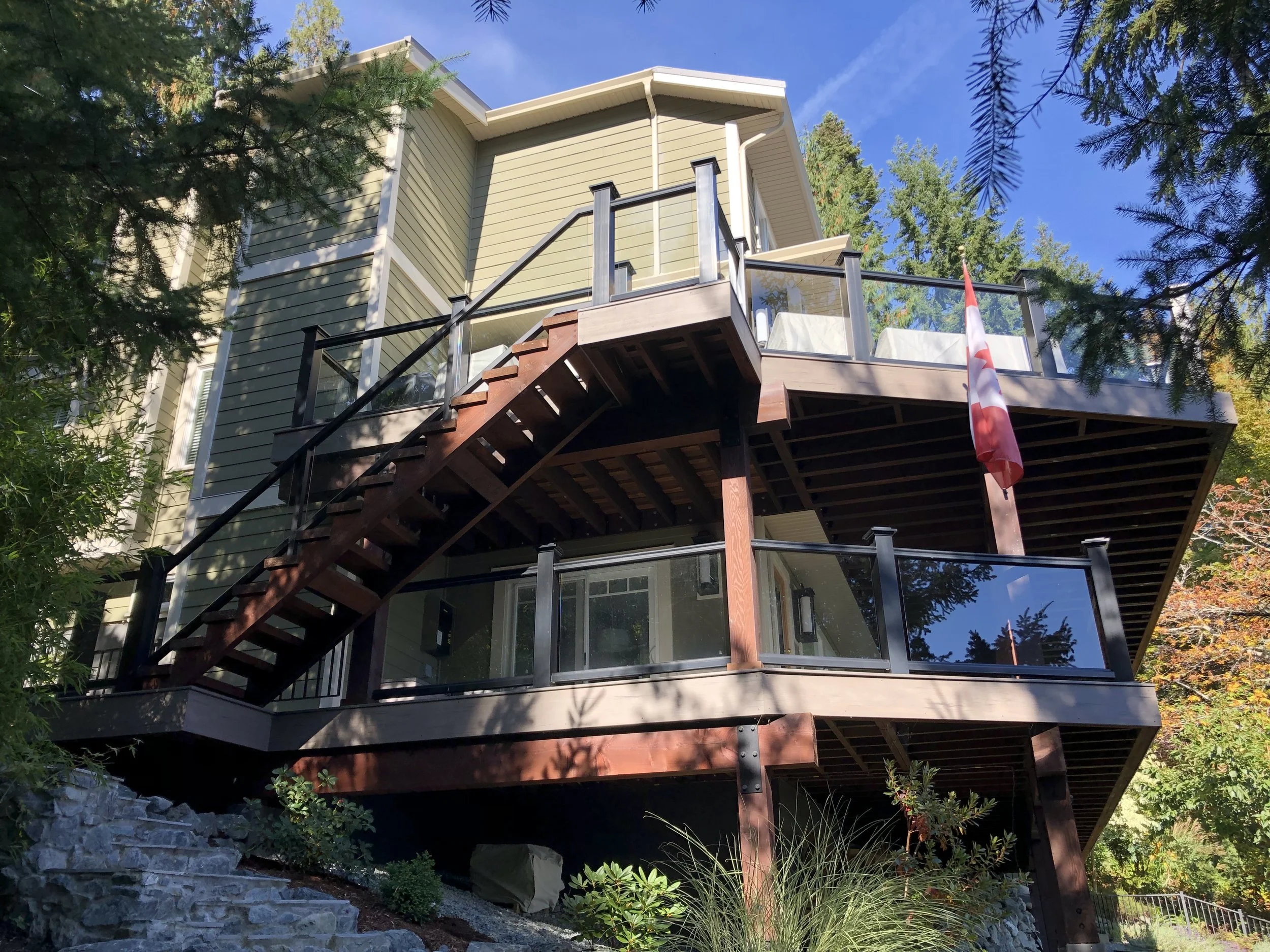 A multi-level house with beige siding, large windows, and a wooden deck enclosed by glass railing. The house is surrounded by tall trees and greenery, with a blue sky overhead.