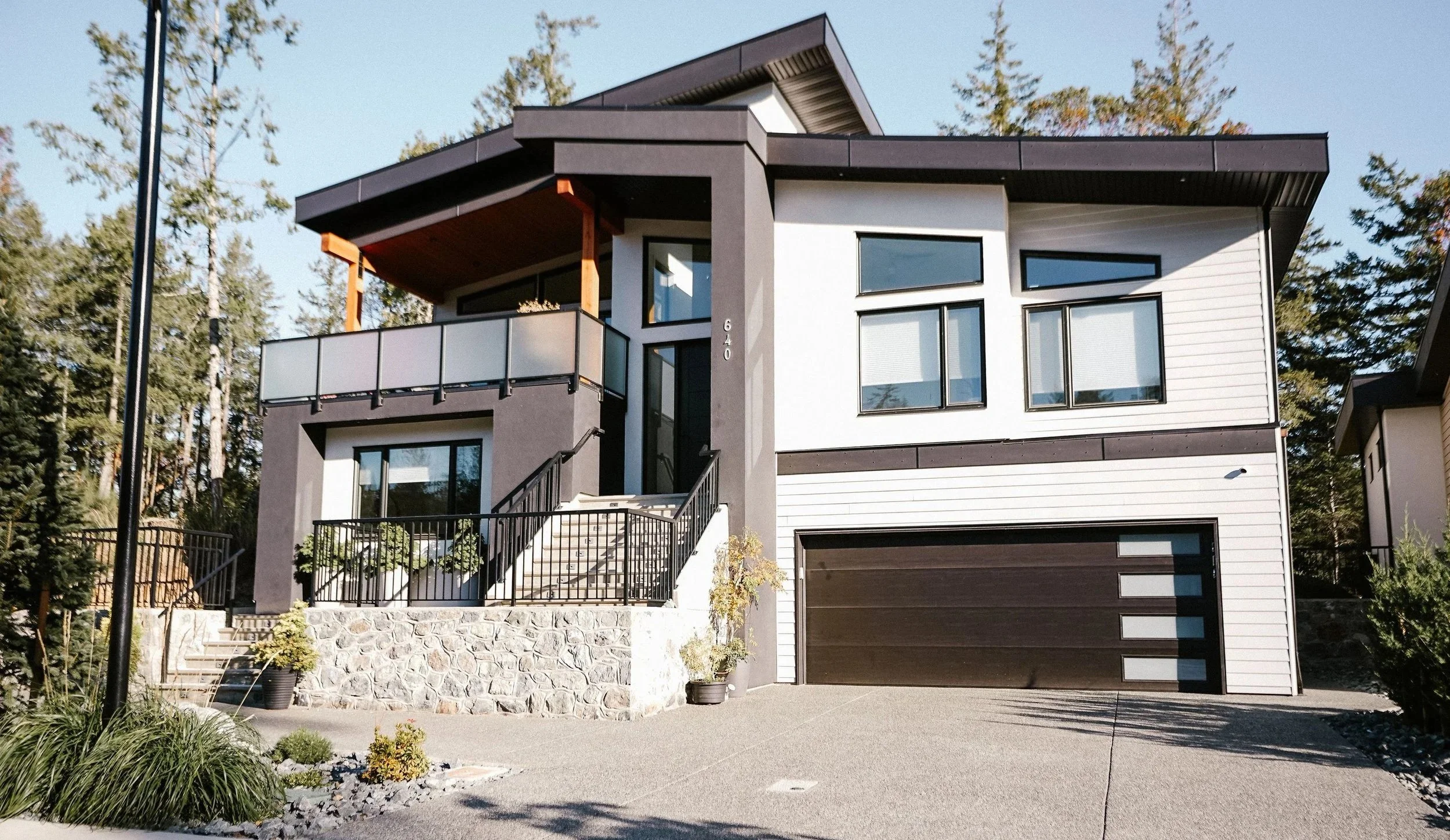 A modern two-story house with black and white exterior, large windows, front steps, and a balcony, surrounded by trees and landscaping.