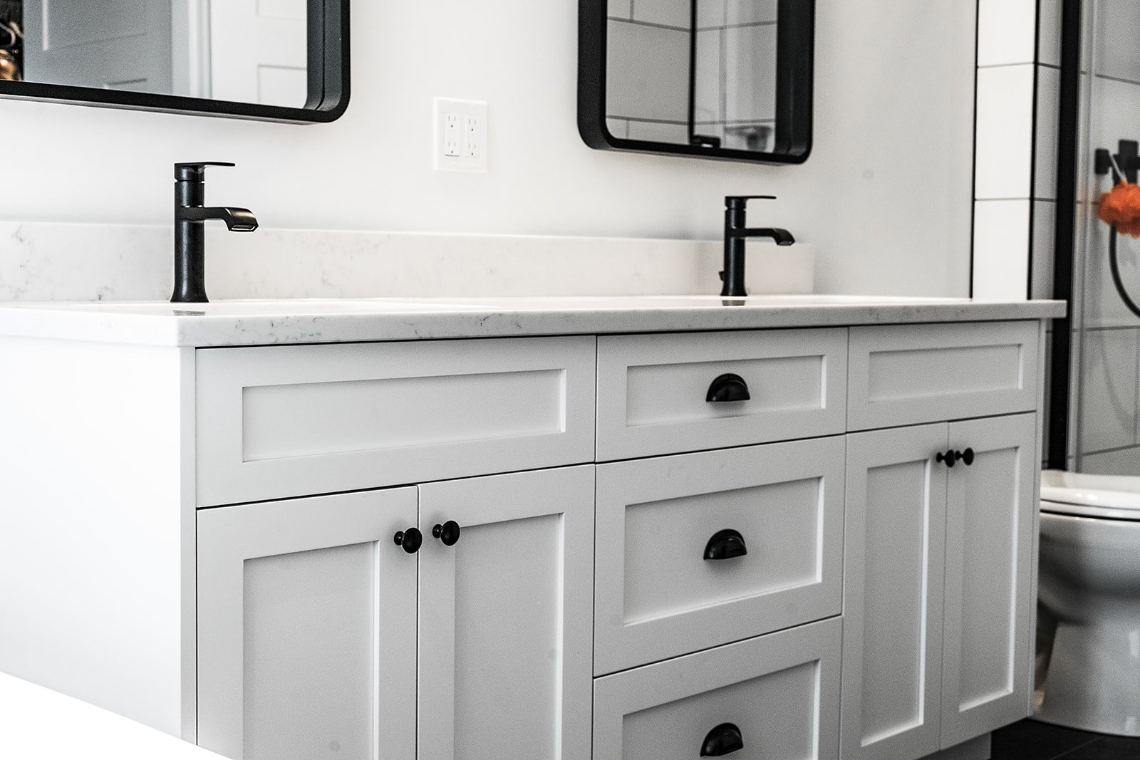 White bathroom vanity with black hardware, double black faucets, mirror, and a glimpse of a toilet in a modern bathroom.