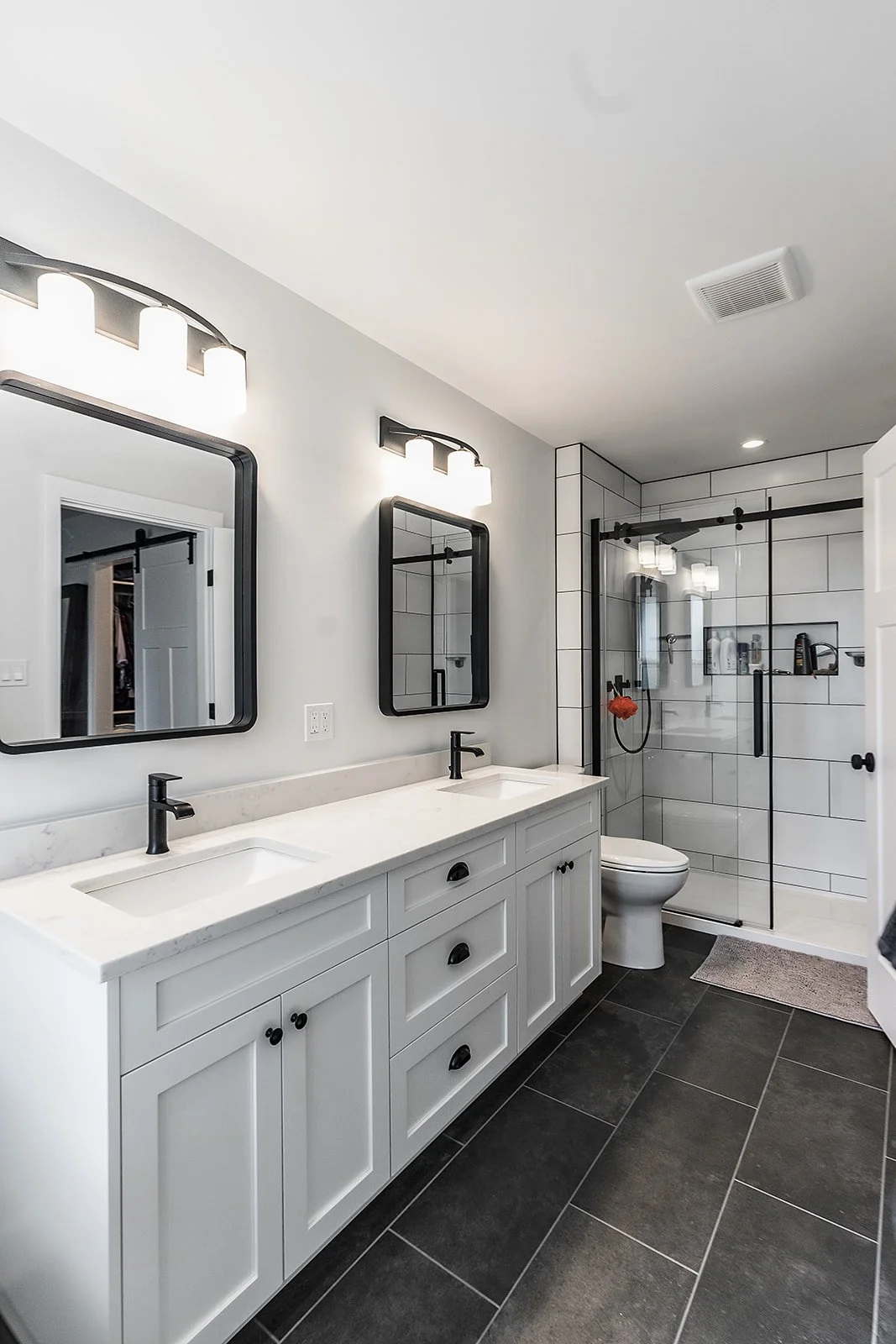 Modern bathroom with double vanity, black framed mirrors, black fixtures, walk-in shower with glass door, white subway tiles, black accents, black tiled floor, ceiling light, and a vent.