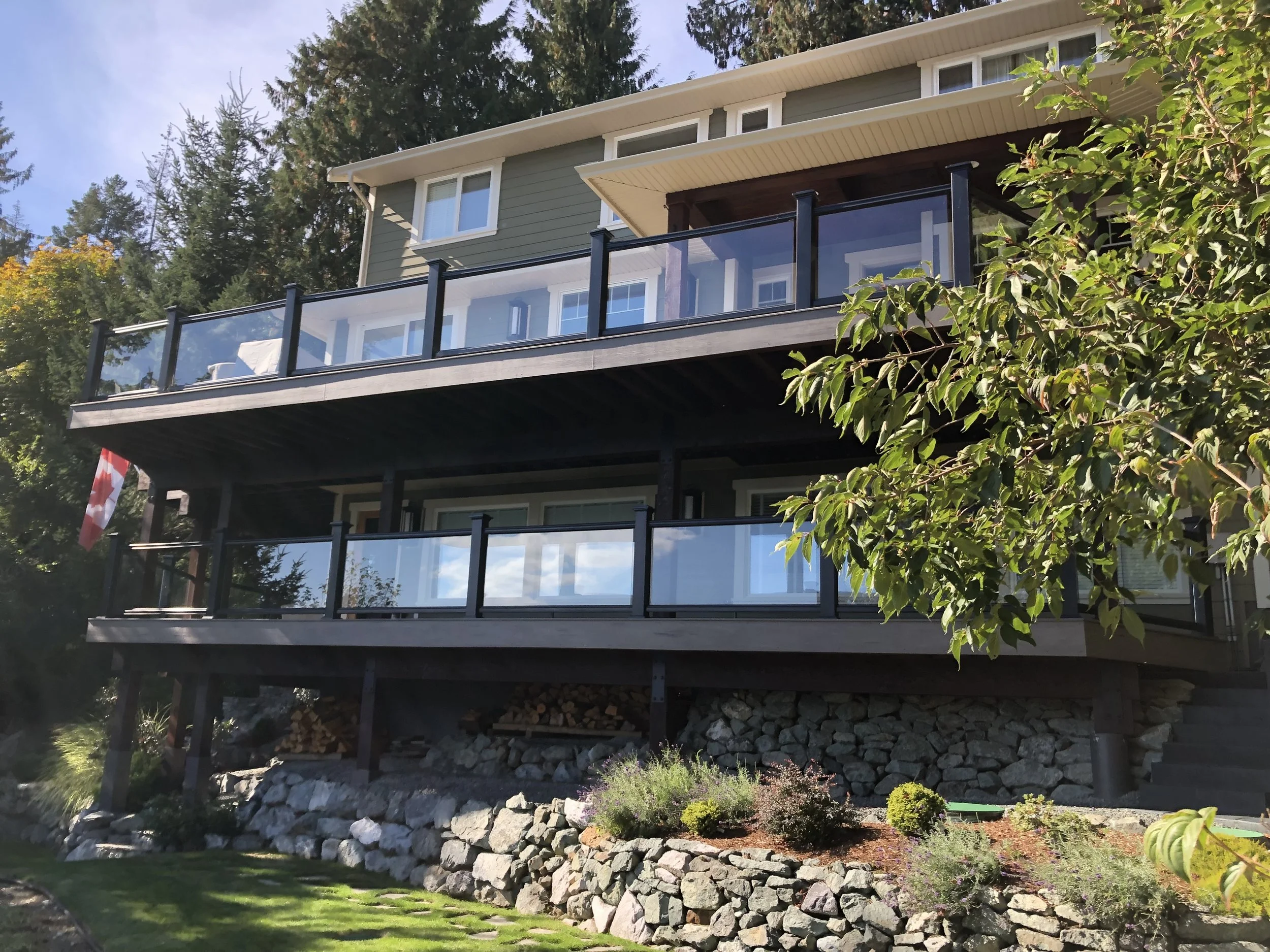 A multi-story house with green and white exterior, large windows, and a spacious balcony with glass railing, surrounded by trees and landscaped garden with rocks and plants.