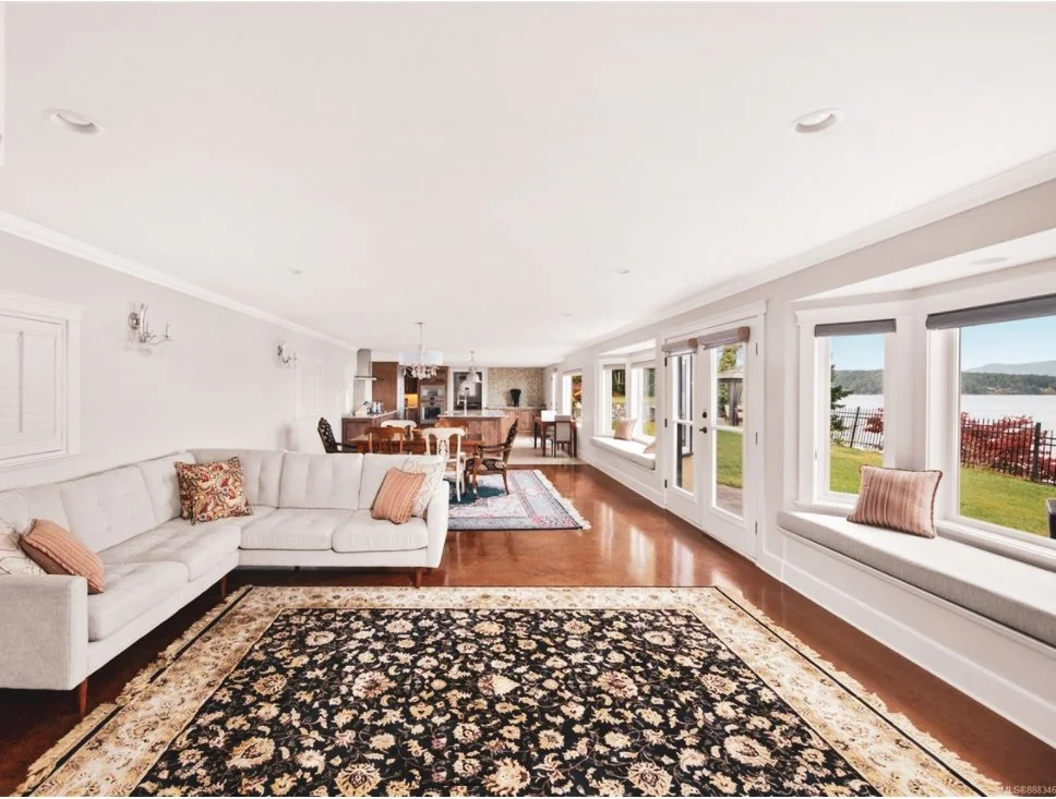Living room with white walls, large white sectional sofa with decorative pillows, floral and beige area rugs, hardwood floors, large windows with views of a lake and backyard, and an open kitchen and dining area in the background.