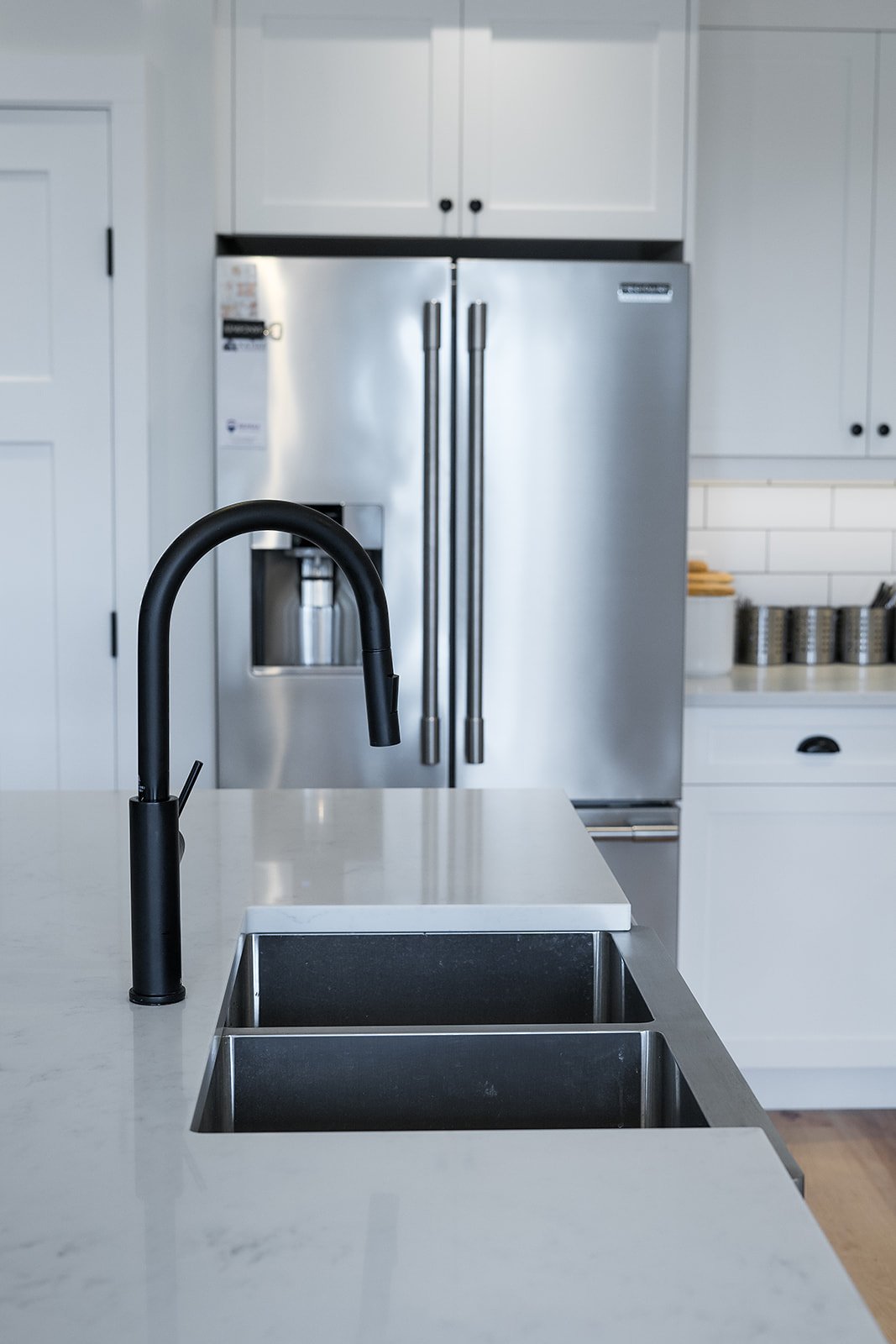 Modern kitchen with a black faucet over a white countertop and stainless steel double sink, with a stainless steel refrigerator in the background.