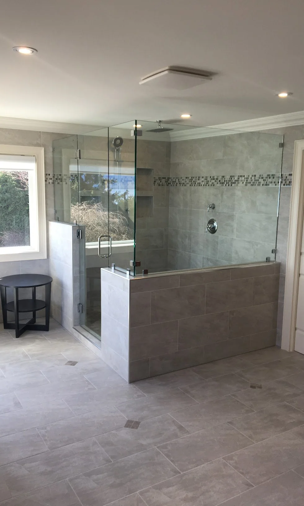 A modern bathroom featuring a spacious walk-in shower with glass enclosure, tiled walls with decorative mosaic strip, a window, and a small black round table.