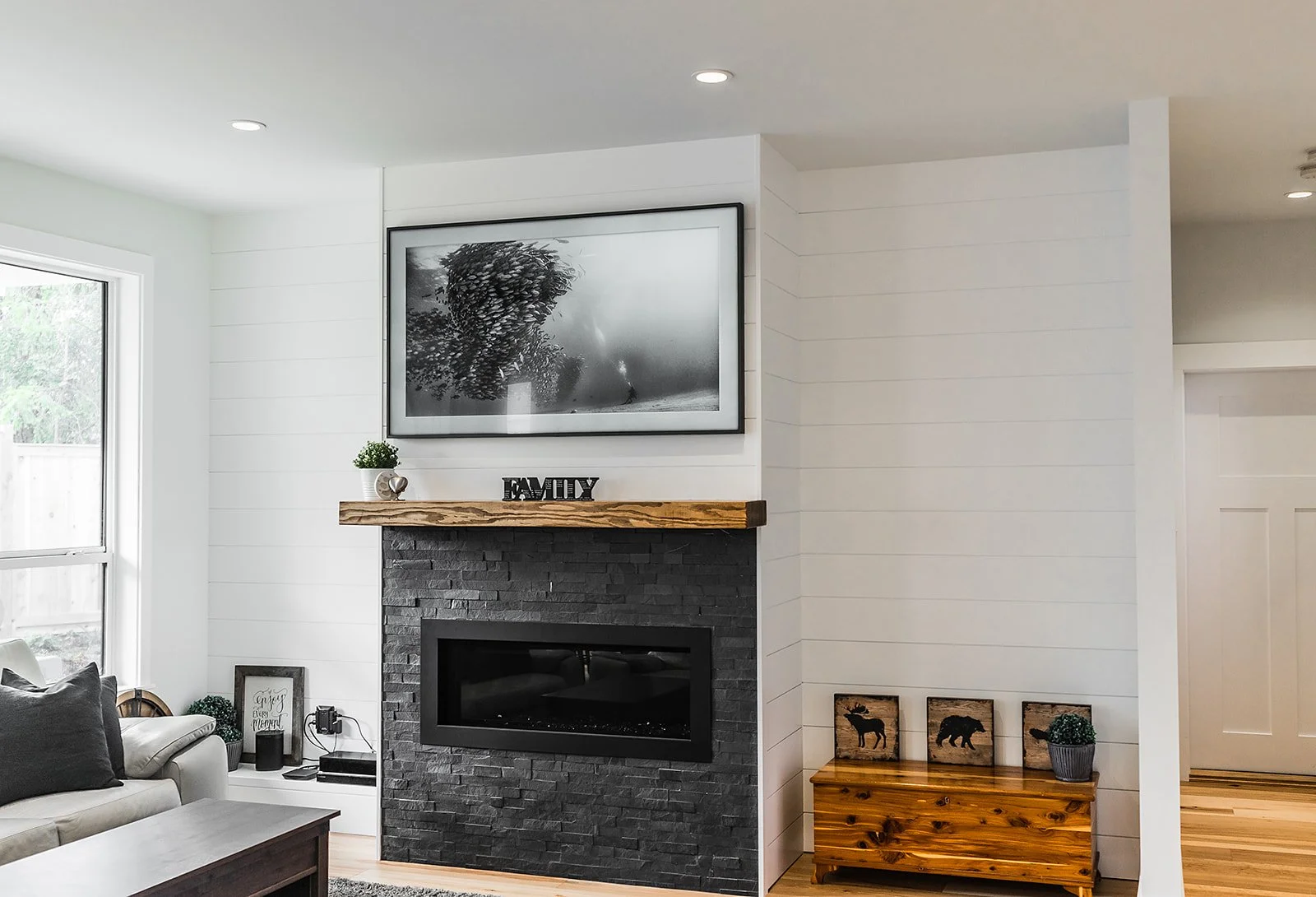 Living room with a black fireplace and a wooden mantel, framed black and white wall art, and decor items including a carved wood sign saying 'FAMILY', small plants, and framed pictures of wildlife.