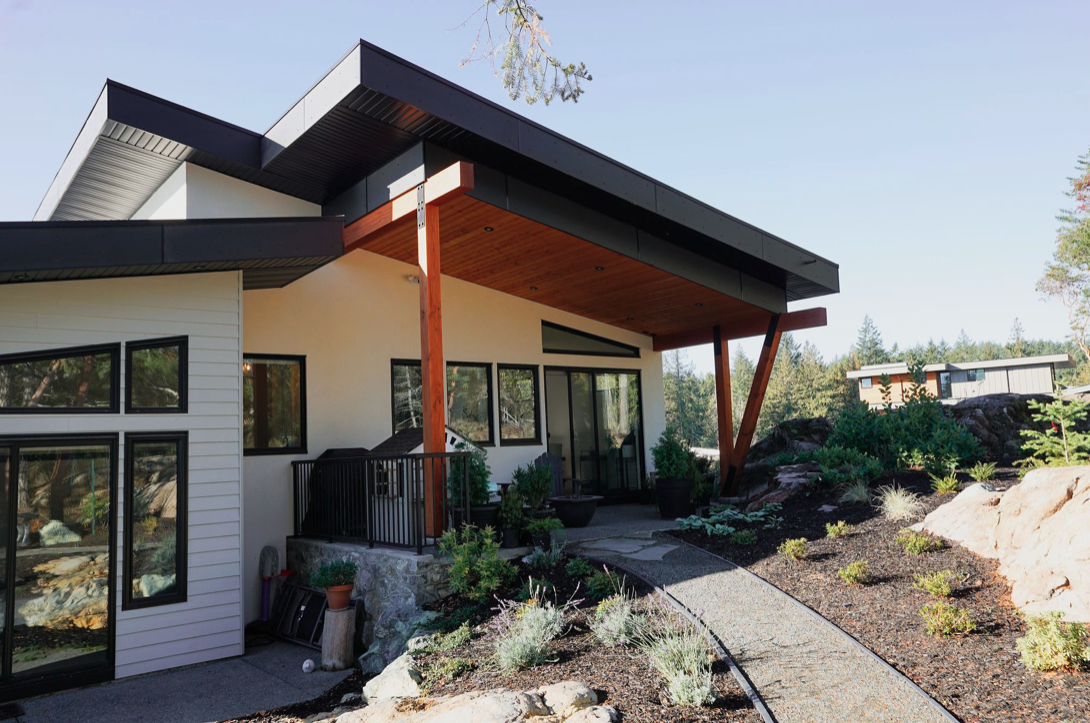 Modern house with white exterior, large black-framed windows, and a wooden patio roof supported by angled wooden beams, surrounded by landscaped garden with rocks and plants.