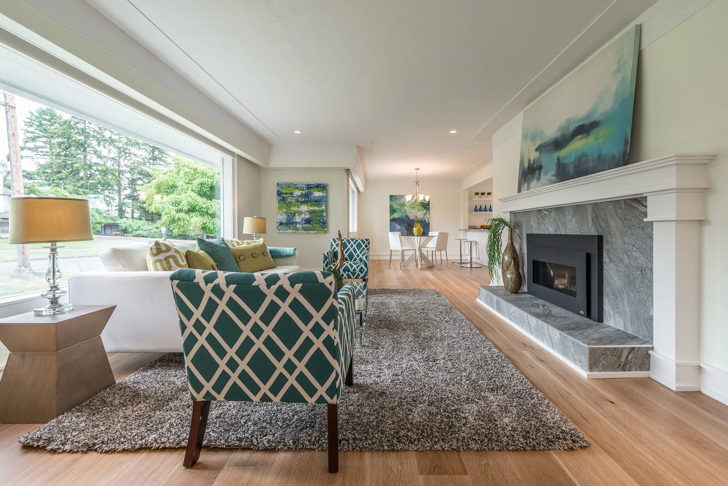 Living room with large window, white sofa, patterned armchair, fireplace, abstract paintings, and hardwood floors.