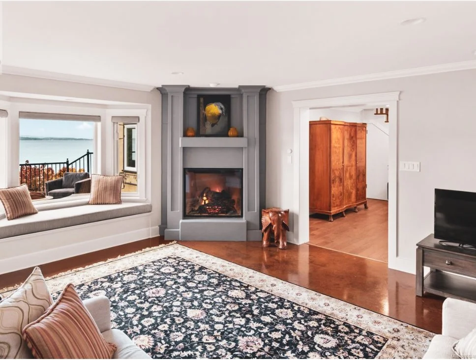 Living room with a fireplace, window seat with cushions, a rug, and a view of water outside, with a wooden cabinet visible through a door frame.