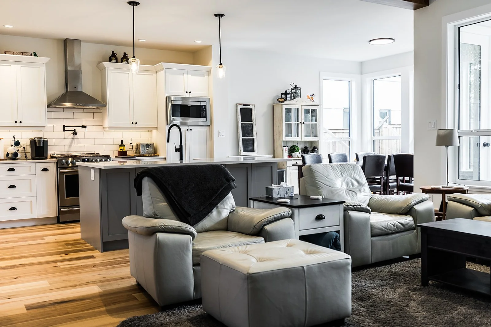 Open-plan living room and kitchen with white cabinetry, a gray kitchen island, beige leather armchairs, a TV console, and large windows letting in natural light.