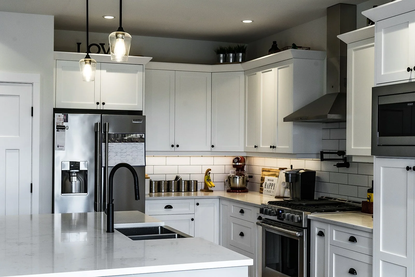 Modern white kitchen with stainless steel refrigerator, black handles, white countertops, black faucet, and various kitchen appliances and decor.