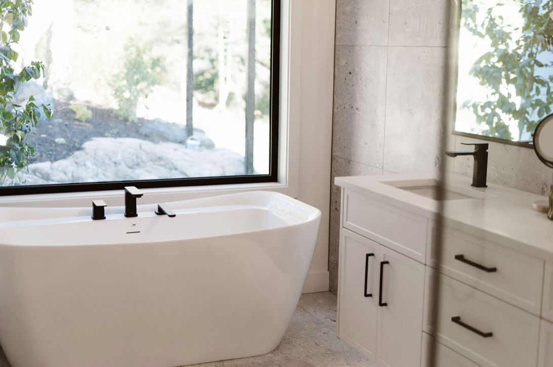 Modern bathroom with a white bathtub near a large window, a white vanity with black handles, and a rectangular mirror.