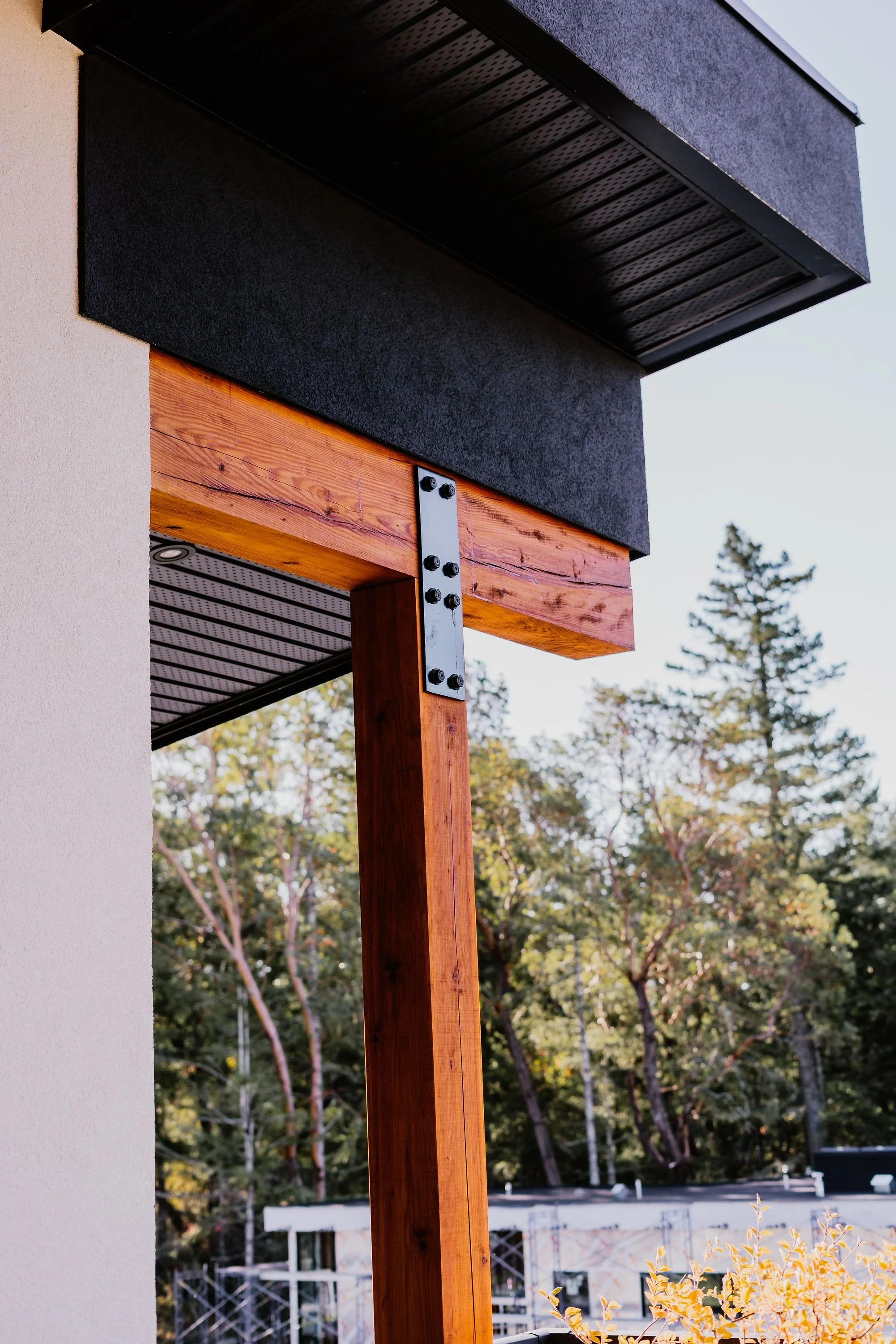 Close-up of a building's exterior corner showing a wooden support beam, black metal bracket, and black textured soffit with a white wall to the left, and trees in the background.