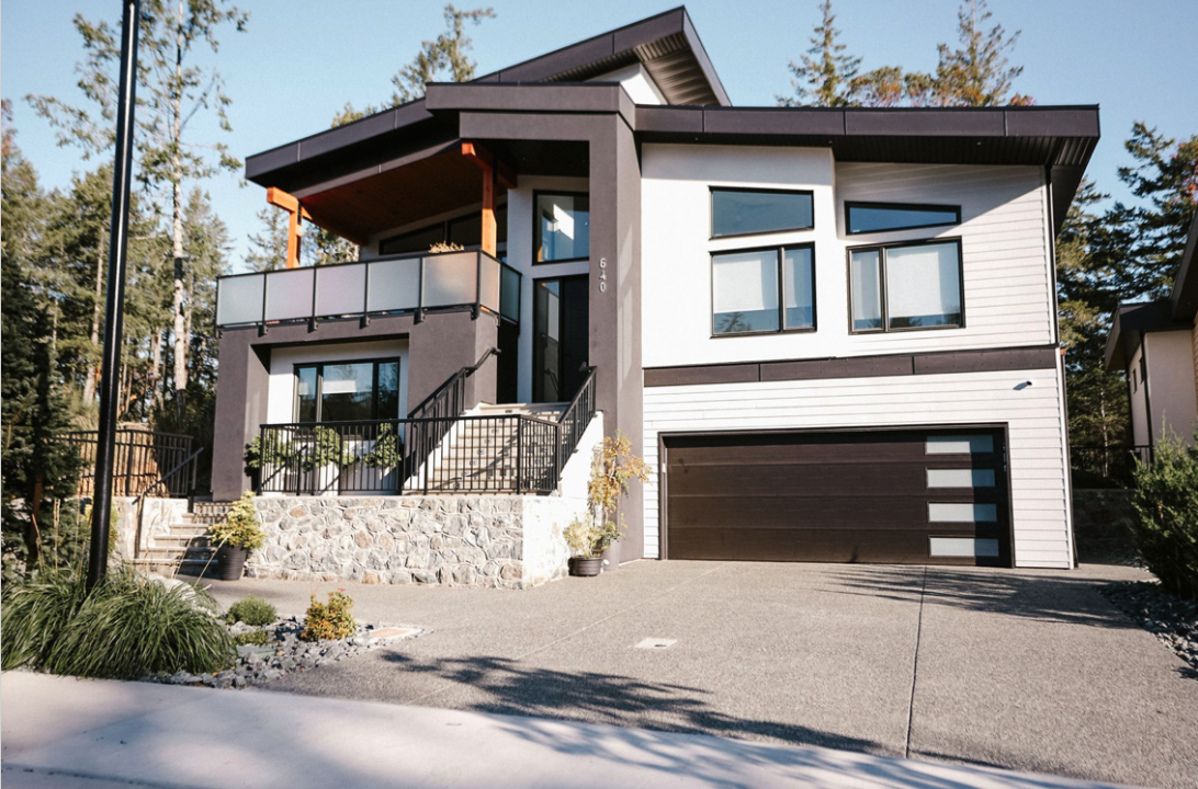 Modern two-story house with a gray and white exterior, large windows, a garage, and a small front yard with plants and trees in the background.