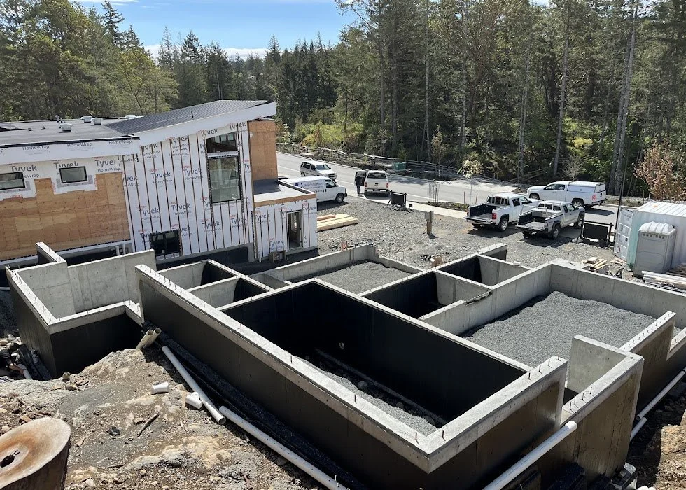 Construction site of a house with visible concrete basement foundation and partially built upper floor framing, surrounded by parked pickup trucks and construction equipment, in a wooded area.