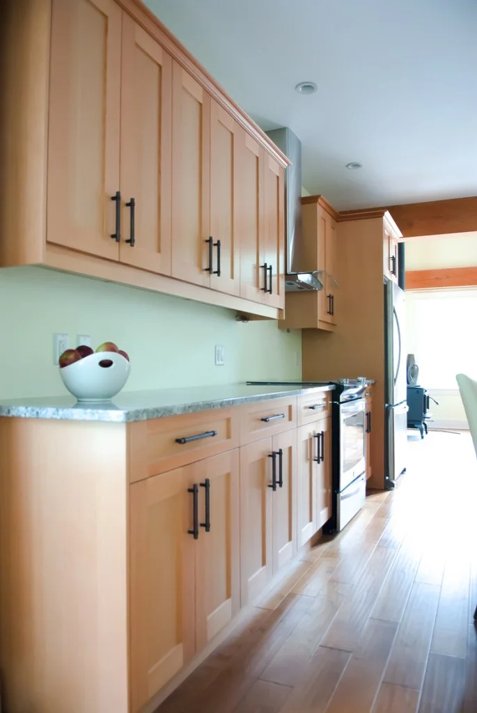 Kitchen with light wood cabinets, marble countertop, and stainless steel appliances, including a refrigerator and oven. A bowl of apples is on the countertop, and sunlight is coming through a window in the background.