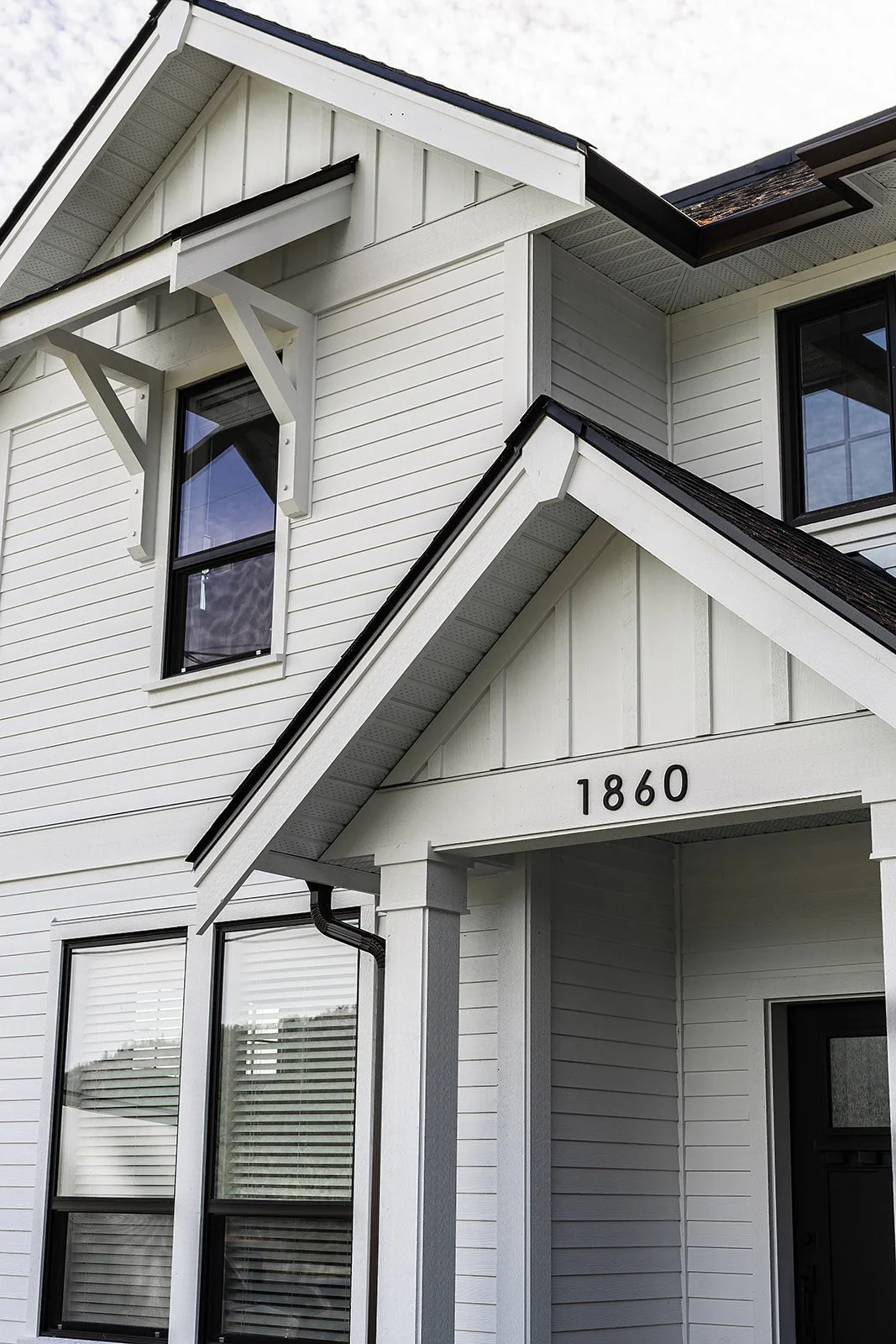 Close-up of a white house with black roof shingles and black window frames, showing a second-story window and part of the front porch with the house number 1860.