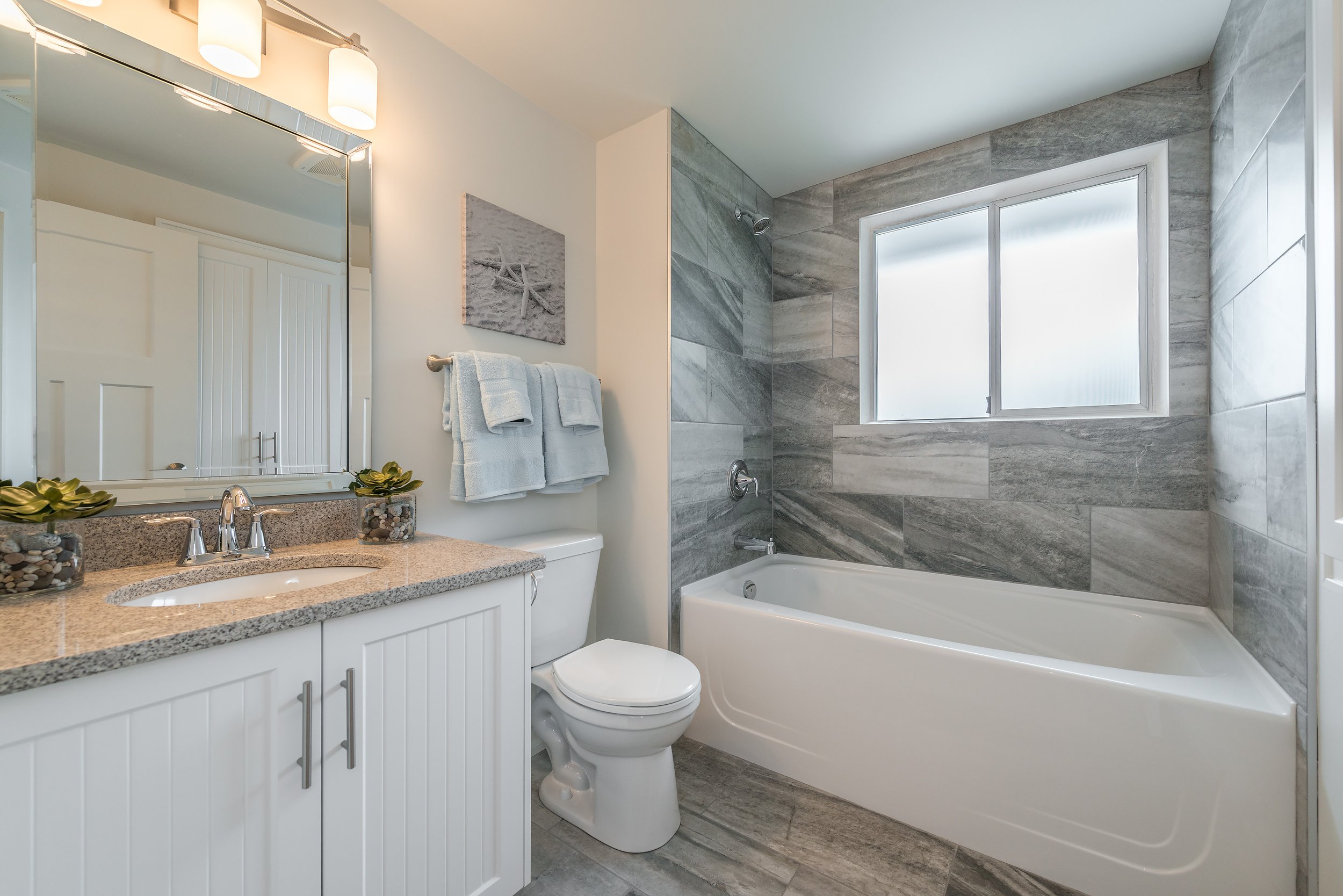 A modern bathroom with a vanity, large mirror, beige granite countertop, white cabinets, a toilet, a bathtub, and a frosted window with gray tile walls.