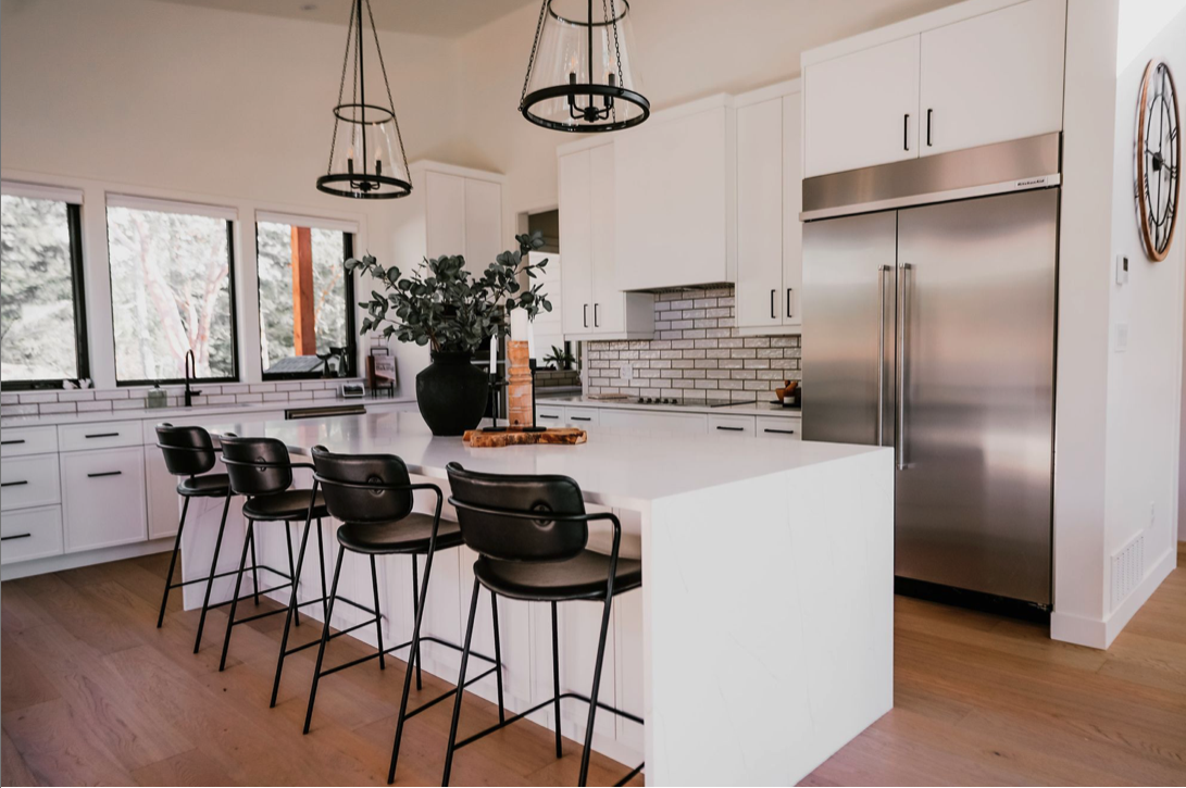 Modern kitchen with white cabinets, stainless steel refrigerator, black bar stools at island, and large windows showing outdoor trees.