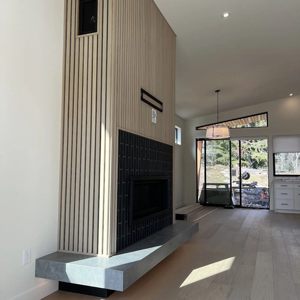 Interior view of a modern living room with a sleek fireplace surrounded by black tiles, a tall wooden slatted wall, and large sliding glass doors leading to an outdoor area. Natural light illuminates the space.
