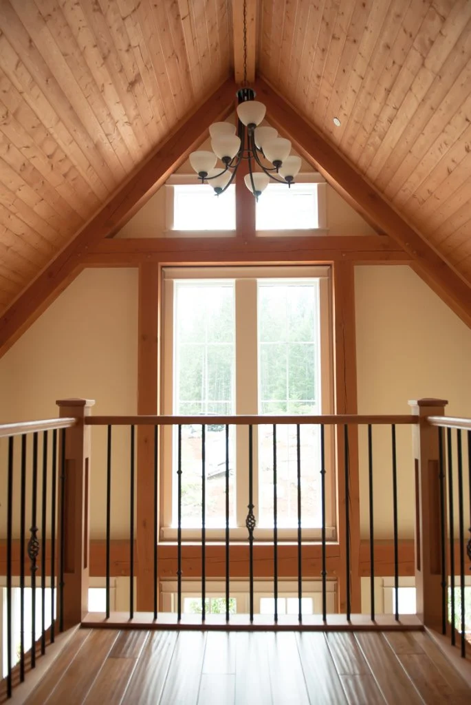 Interior view of a room with a large window featuring a wooden frame and mullions, high vaulted wood-paneled ceiling, and a chandelier with multiple lamps hanging from the ceiling.
