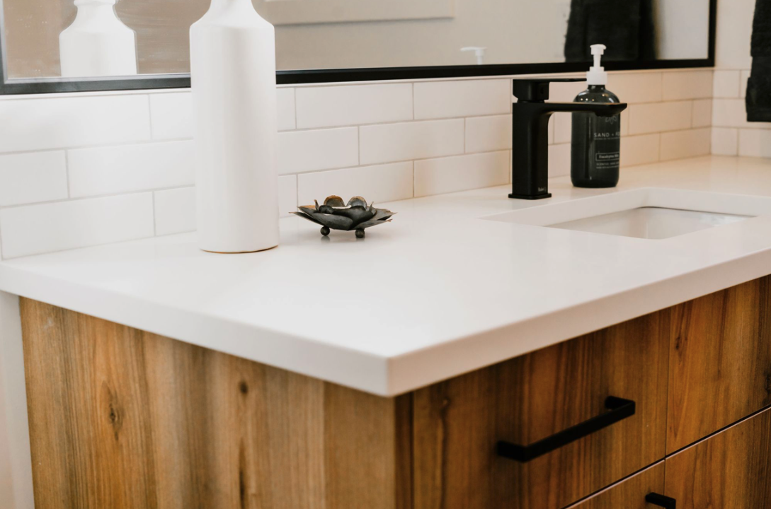 Bathroom vanity with white countertop, black faucet, soap dispenser, decorative dish with small objects, and a mirror.