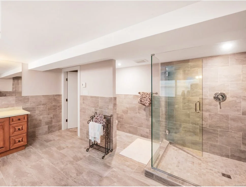 A modern bathroom with beige tiled walls and floor, a glass-enclosed shower, a wooden cabinet on the left, and a towel rack with towels.