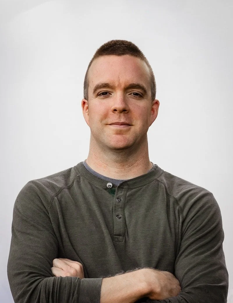 Portrait of a man with short hair, crossing his arms, wearing a dark gray long-sleeve shirt, standing against a plain light gray background.