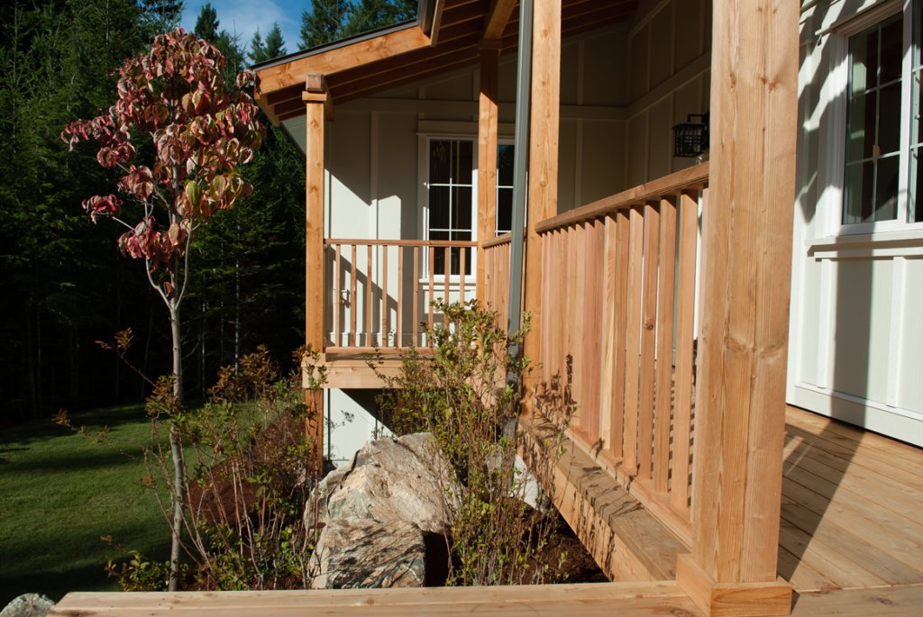 A wooden porch with a railing attached to a house, surrounded by plants and a small tree, with a forest in the background.