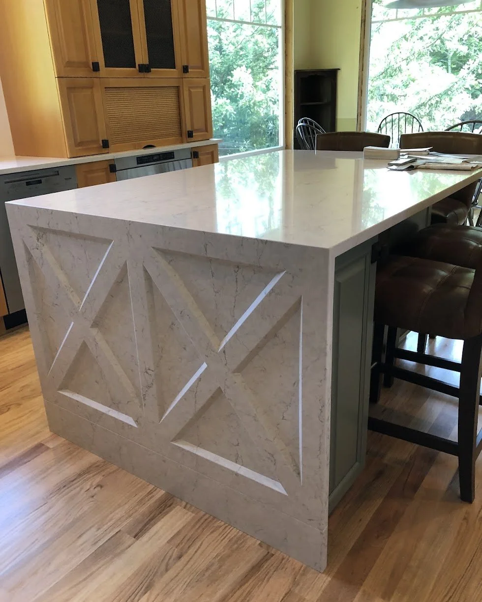 A kitchen island with a marble top and decorated sides, surrounded by chairs at a breakfast bar, with a wooden cabinet and view of trees through large windows in the background.
