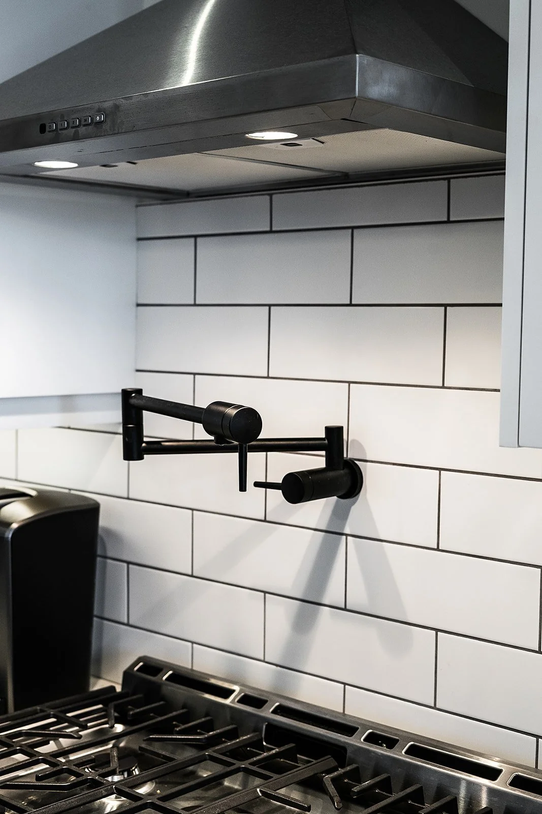 Close-up of a modern kitchen with a stainless steel range hood, white subway tile backsplash, and black wall-mounted pot fillers above a gas stove.